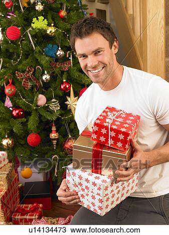 Stock Image - Young man holding gifts in front of Christmas tree. Fotosearch