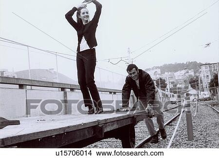 Picture - Young woman standing on a trolley being pushed by a young man on a railroad track. Fotosearch