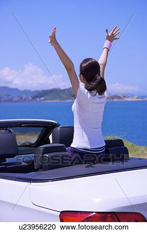Stock Image - Young woman sitting in the car with arms outstretched and looking up. Fotosearch
