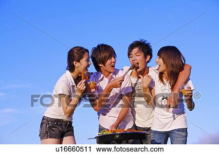 Stock Image - Four people barbecuing outside and eating food,. Fotosearch