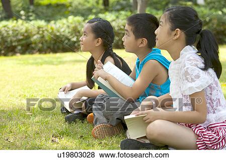 Stock Photo - Three children sitting on the lawn with books and listening together. Fotosearch
