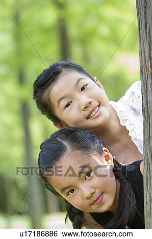 Stock Photograph - Two girls hiding behind a tree together and looking at the camera. Fotosearch
