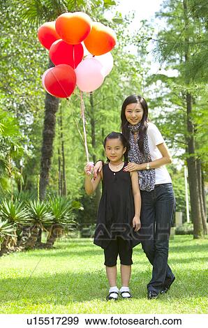 Young woman and girl standing on the lawn together and holding balloon View Large Photo Image Stock Photo - Young woman and girl standing on the lawn together and holding balloon. Fotosearch