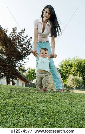 Stock Photo - Young woman helping baby boy walk in garden. Fotosearch