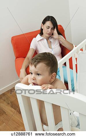 Stock Image - Young woman on arm chair holding her head and baby boy in crib. Fotosearch
