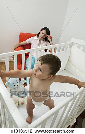 Stock Image - Young woman on arm chair holding her head and baby boy in crib. Fotosearch