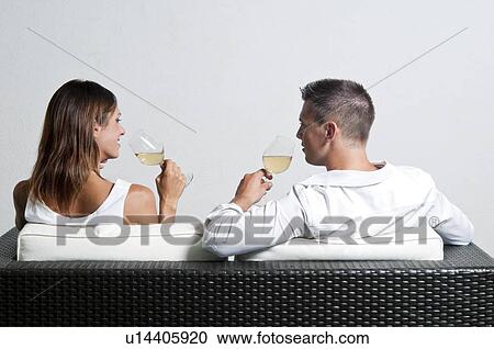 Stock Image - View of couple from the back sitting on couch drinking white wine. Fotosearch