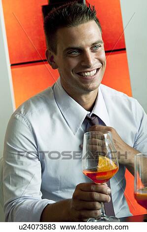 Stock Image - Young man in business attire with glass of orange wine adjusting tie. Fotosearch