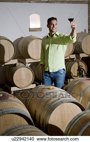 Stock Image - Young man in wine cellar holding up glass of red wine. Fotosearch
