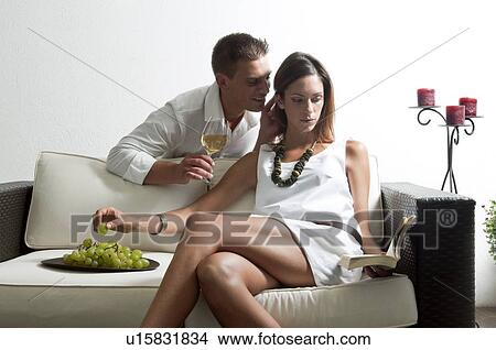 Picture - Young man leaning on couch with white wine whispering in young woman's ear reading book and eating grapes. Fotosearch