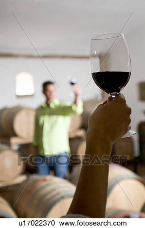 Stock Image - Young woman's hand holding glass of red wine with young man in background holding up his wineglass. Fotosearch