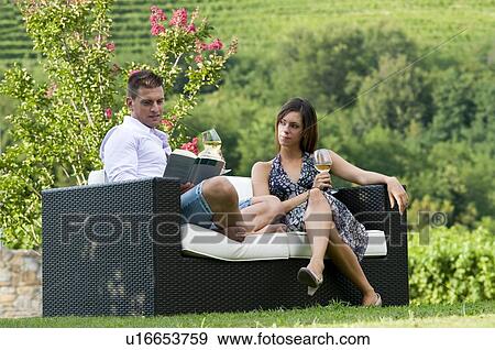 Stock Photo - Young couple sitting on couch in vineyard with white wine, man reading book. Fotosearch