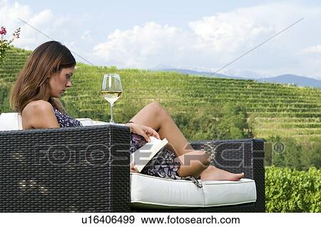 Stock Photo - Young woman sitting on couch in vineyard with white wine reading a book. Fotosearch