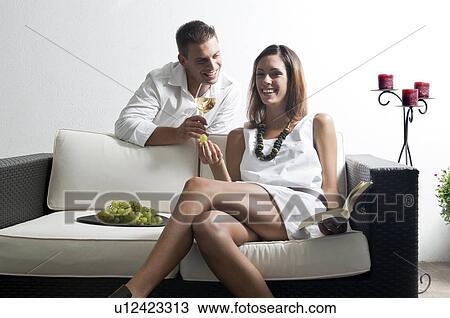 Stock Image - Young man leaning on couch with white wine and young woman reading book and eating grapes. Fotosearch