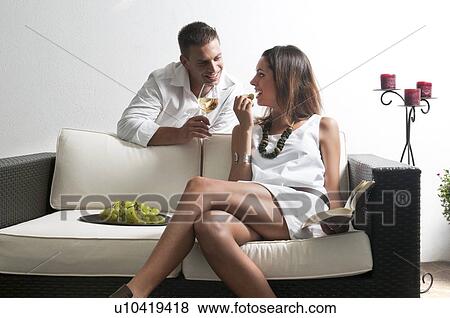 Stock Photo - Young man leaning on couch with white wine and young woman reading book and eating grapes. Fotosearch