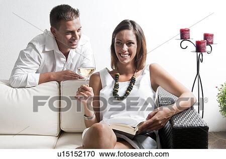 Stock Image - Young man leaning on couch with young woman with book and white wine. Fotosearch