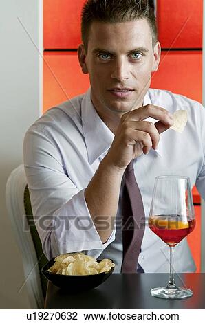 Stock Image - Young man sitting with a glass of orange wine and potato chips. Fotosearch