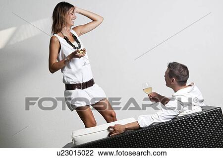 Stock Image - Young woman with white wine standing next to couch with young man and wine. Fotosearch