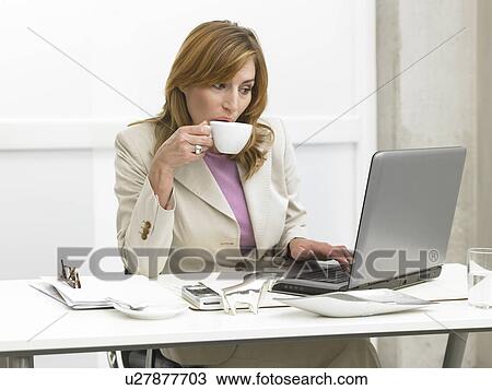 Stock Image - Business woman is drinking coffee at her desk. Fotosearch