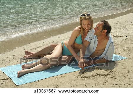 Stock Photo - Couple lying on the beach, he is painting a heart into the sand. Fotosearch