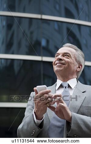 Picture - Senior businessman holding mobile phone and looking up with smile. Fotosearch