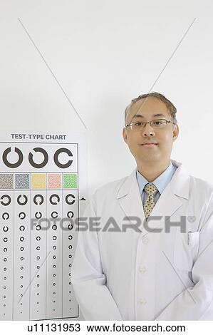 Stock Image - Doctor standing before eye chart and looking at the camera. Fotosearch