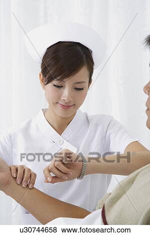 Stock Photo - Young nurse taking pulse for the patient and looking at the watch. Fotosearch