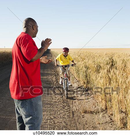 Stock Image - Father clapping as son (8-9) is cycling along dirt road in fields. Fotosearch