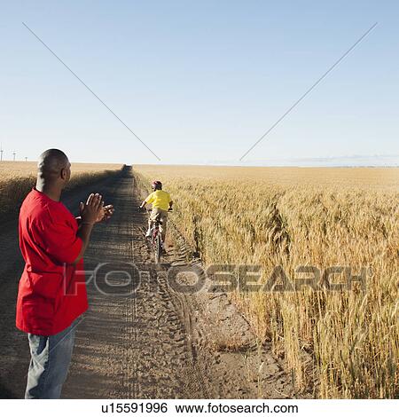 Stock Photograph - Father clapping as son (8-9) is cycling along dirt road in fields. Fotosearch