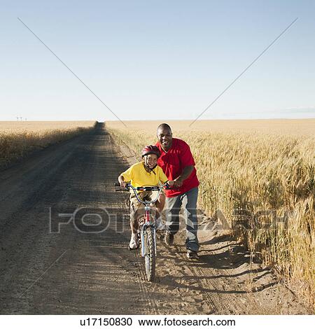 Stock Image - Father teaching son (8-9) how to cycle on along dirt road. Fotosearch