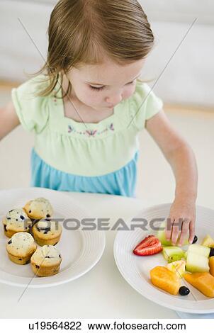 Stock Image - Girl (2-3) choosing between fruit salad and muffins. Fotosearch