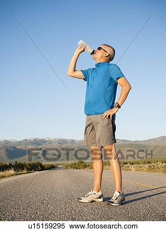 Stock Photography - Mid adult an drinking water while taking break from running on empty road. Fotosearch