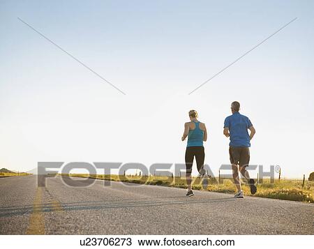 Stock Image - Mid adult couple running on empty road. Fotosearch