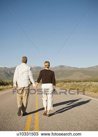 Stock Image - Mid adult couple walking along empty road . Fotosearch