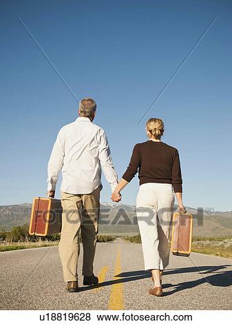 Stock Photo - Mid adult couple walking along empty road carrying suitcases. Fotosearch