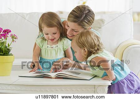 Stock Image - Mother with daughters (2-3) reading book in living room. Fotosearch
