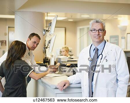Stock Image - Portrait of confident doctor with two colleagues working in background. Fotosearch