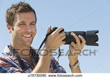 Portrait of man holding camera against blue sky View Large Photo Image Stock Image - Portrait of man holding camera against blue sky. Fotosearch