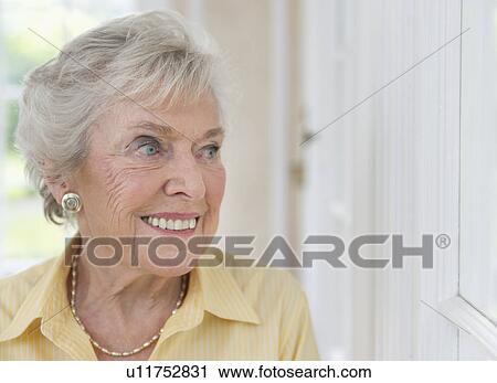 Stock Image - Portrait of senior woman looking through window. Fotosearch