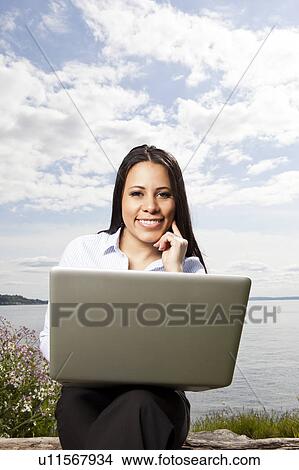 Picture - Portrait of young businesswoman using laptop on lakeshore. Fotosearch