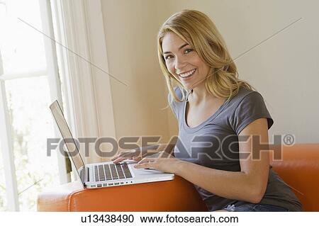 Stock Image - Portrait of young woman sitting on sofa using laptop. Fotosearch