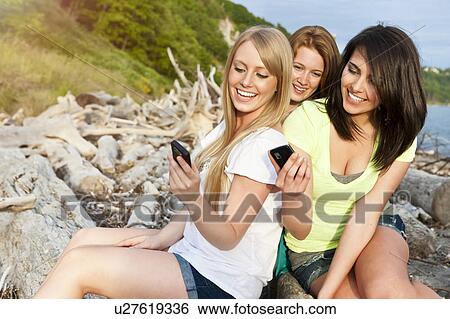 Stock Photograph - Three young women hanging out on beach, using cell phones. Fotosearch