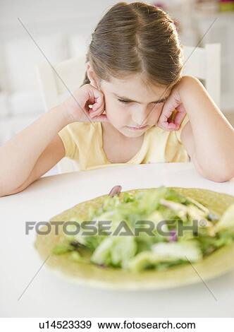 Stock Photo - Upset Girl ( 6-7) sitting at table with salad meal. Fotosearch
