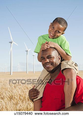 Stock Image - USA, Oregon, Wasco, Boy (8-9) piggy-back riding on father, wind turbines in background. Fotosearch