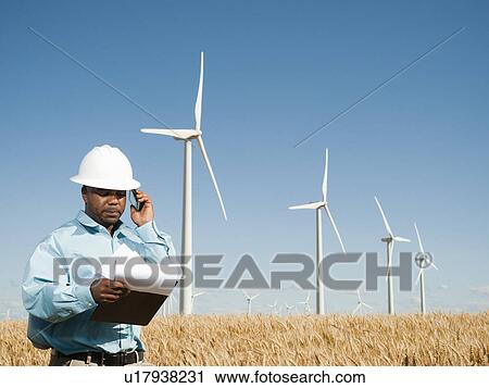 Stock Image - USA, Oregon, Wasco, Engineer standing in wheat field in front of wind turbines. Fotosearch
