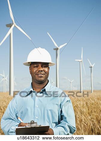 Stock Image - USA, Oregon, Wasco, Engineer standing in wheat field in front of wind turbines. Fotosearch
