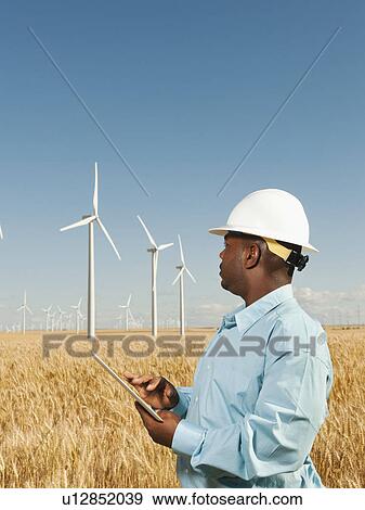 Stock Photo - USA, Oregon, Wasco, Engineer standing in wheat field in front of wind turbines, using digital tablet. Fotosearch