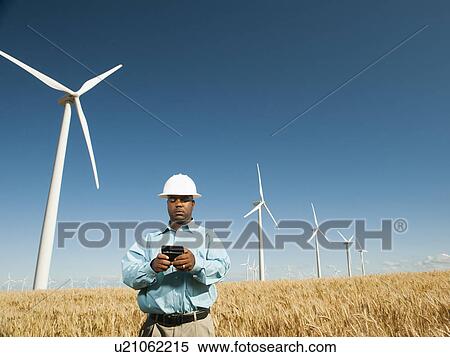 Stock Photography - USA, Oregon, Wasco, Engineer standing in wheat field in front of wind turbines, using mobile phone. Fotosearch