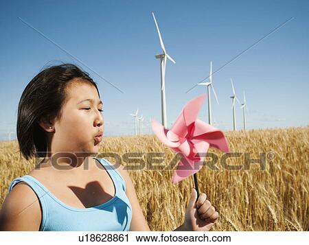 Stock Image - USA, Oregon, Wasco, Girl (10-11) holding blowing at fan in wheat field with wind turbines in background. Fotosearch