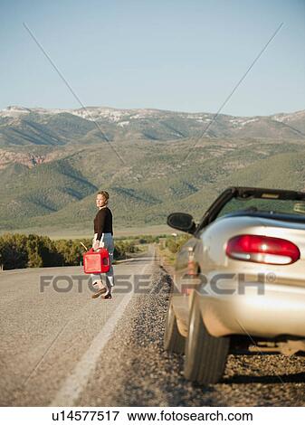 Stock Photo - USA, Utah, Kanosh, Woman carrying canister walking along empty road, her car parked on roadside. Fotosearch